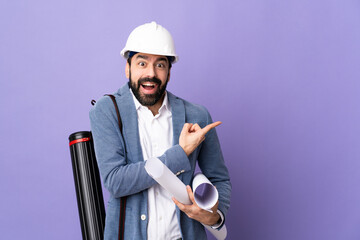 Young architect man with helmet and holding blueprints over isolated purple background surprised...