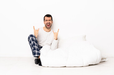 Caucasian man in pajamas sitting on the floor at indoors making horn gesture