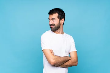 Tuinposter Young man with beard  over isolated blue background with arms crossed and happy © luismolinero