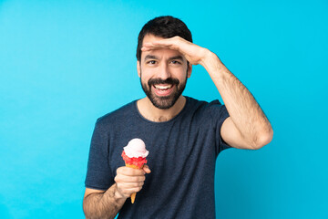 Young man with a cornet ice cream over isolated blue background looking far away with hand to look...