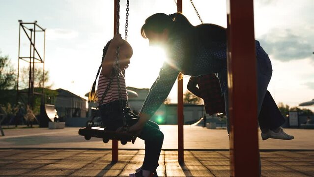 Caring young mother swinging on swing cute little daughter girl at sunny summer outdoor sport playground. Happy woman enjoy motherhood spending time with kid happy childhood leisure activity