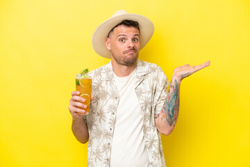 Young caucasian man holding a cocktail isolated on yellow background having doubts while raising hands - obrazy, fototapety, plakaty
