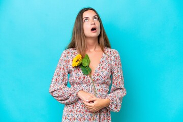 Young caucasian woman holding sunflower isolated on blue background looking up and with surprised...