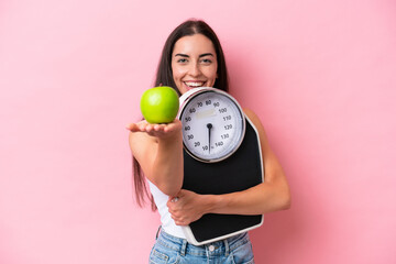 Young caucasian woman isolated on pink background holding weighing machine and offering an apple