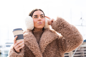 Young woman wearing winter muffs  and holding take away coffee at outdoors having doubts and with...