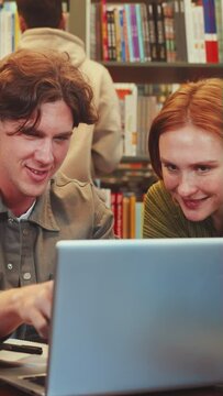 Vertical screen: Two focused students engage in collaborative study using laptop at library table, surrounded by shelves packed with colorful books. Another persons browse books in background