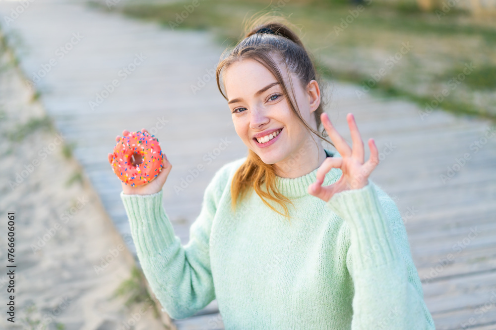 Wall mural Young pretty girl holding a donut at outdoors showing ok sign with fingers