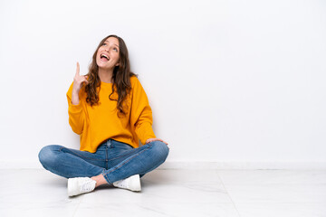 Young caucasian woman sitting on the floor isolated on white wall pointing up and surprised