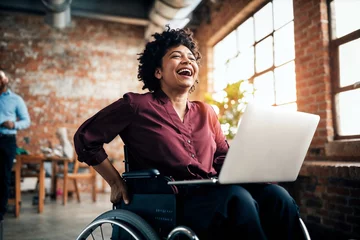 Foto auf Leinwand African American businesswoman in a wheelchair laughs while working on her laptop in a creative office space, embodying success and accessibility.   © InputUX
