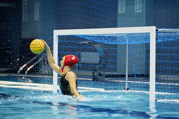 Young active woman in swimming cap and swimsuit catching ball of rival team while standing on gate defense during water polo game