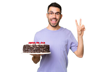Young caucasian man holding birthday cake isolated on green chrome background smiling and showing victory sign