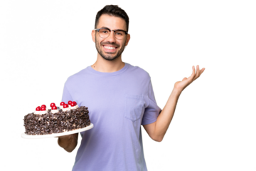 Young caucasian man holding birthday cake isolated on green chrome background extending hands to the side for inviting to come