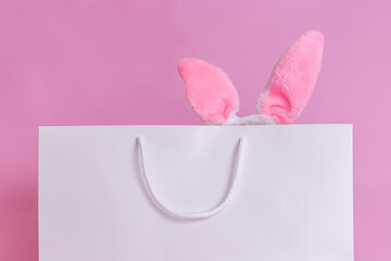 Close-up of a white paper bag on a pink background, with fluffy pink Easter bunny ears sticking out.