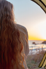 woman with long hair in a tent meets the sunrise on the sea coast, rear view. Travel and tourism