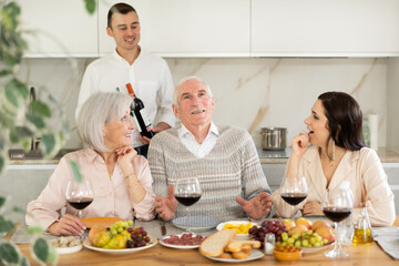 Glad mature man and woman, their son and daughter-in-low sitting at table while having friendly conversation with a glass of wine at home