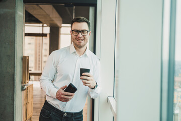 Naklejka na ściany i meble A male manager in a white shirt and glasses stands in the office by the window and drinks coffee while watching a video on the phone. Business telephone conversation of a business man.
