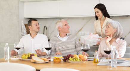 Young lady handing a gift to senior male family member sitting around table and drinking wine together at home
