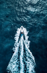 Vue aérienne d'un bateau à moteur blanc se déplaçant dans une eau bleue foncé avec une traînée d'écume blanche  - obrazy, fototapety, plakaty