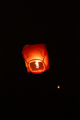 A single glowing orange sky lantern rises against a dark night background, surrounded by faint...