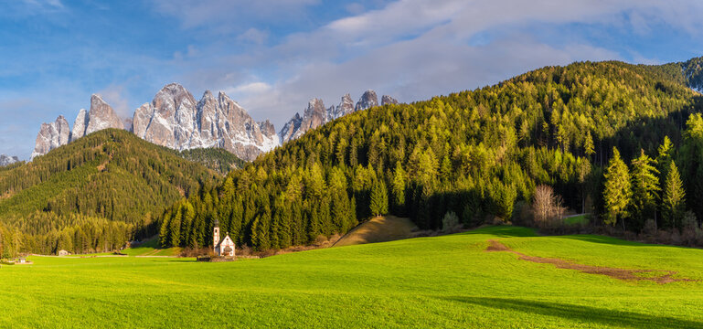Panoramic view of sunny landscape with church of St. John against the Geisler mountains covered with snow in Santa Maddalena village, Val Di Funes in Dolomites mountains, South Tyrol, Italy