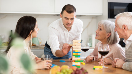 Mature parents and son with girlfriend are having fun, playing building tower with wooden blocks game. Participant is removing wooden block bricks from tower and others are watching.