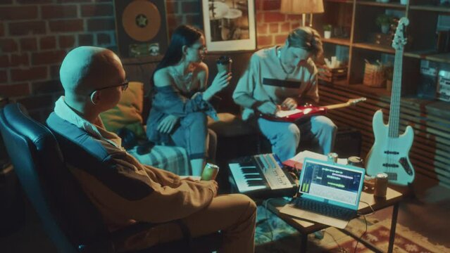 Back footage of musician listening to friends in process creating lyrics for new rock song while sitting together in vintage studio in evening
