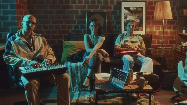 Pull out portrait of Caucasian indie rock group posing on sofa and chair in vintage studio with electric guitar and midi controller and looking at camera