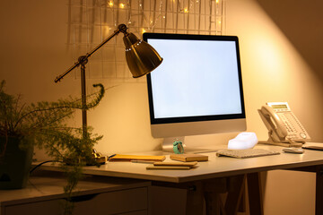 Blank computer monitor with stationery and glowing lamp in office at night