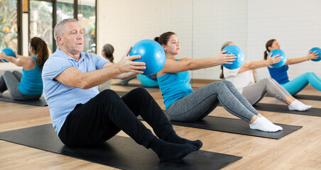 Fototapeta premium Portrait of active senior man during group core training, sitting on mat and doing sit-ups with bender ball at gym. Healthy lifestyle and pilates concept