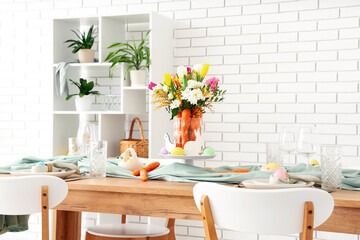 Beautiful table setting for Easter decorated with bouquet of carrots and flowers near white brick wall
