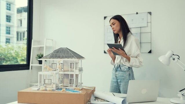 Beautiful caucasian architect focus on using tablet to check house model on meeting table with blueprint, house model and architectural document scatter around at meeting room. Immaculate.
