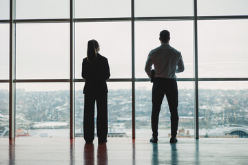 A man and a woman are standing in the office against the background of a large panoramic window. Modern spacious workspace with employees. Business partners