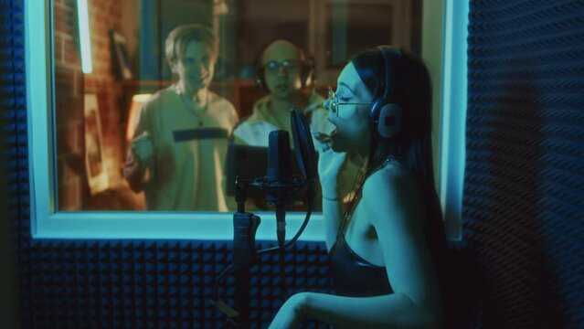 Side footage of Caucasian female vocalist in headset signing into microphone in recording booth while music producer controlling recording in studio