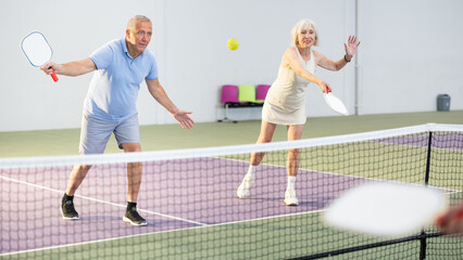 Focused positive aged couple, man and woman, playing friendly doubles pickleball match on indoors court. Healthy active lifestyle of elderly..