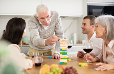 Joyful senior parents and young couple sitting around table playing brick game and drinking wine in goblets at home