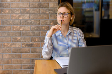 The woman in the office is engaged in business, thought, works on a laptop. Beautiful young woman with glasses and blue shirt, at work or student, freelancer. A woman is engaged on a laptop