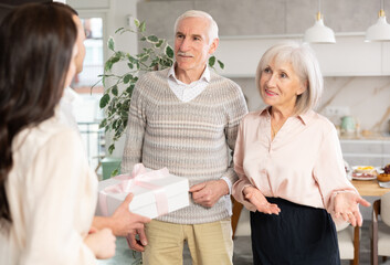 Hospitable smiling elderly couple warmly welcoming adult daughter with husband bringing gift to...