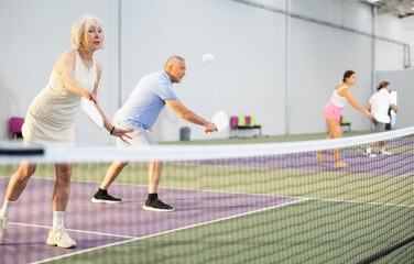 Portrait of sporty senior woman playing doubles pickleball with male partner on indoor court, ready...