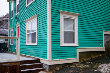 The corner view of an exterior teal green wooden clapboard covered historic building. There are multiple glass windows with white trim. The front door is red with a wood patio and three steps.