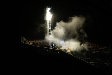 The front of a large ship at night with lights on the bow exposing thick smoke, mist, and bad...
