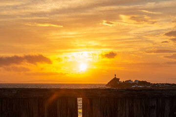 A level wooden breakwater near a beach with a vibrant orange sky and clouds in the background. The wall is made of logs with the bark on the outside. You can see ocean in the spaces through the logs. - obrazy, fototapety, plakaty