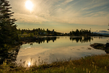 A bright orange sunset over a peaceful and calm river. The evening sky has wispy clouds. Tall evergreen trees surround the water and reflect in the tranquil pond.