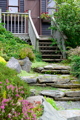 The entrance to a quaint garden shed with a wooden rail and wood steps. The brown building has a rock garden in front. The flowers are yellow, pink, and purple. The plants and trees are lush green.