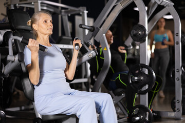 Concentrated sporty senior woman in blue activewear working out shoulders muscles on exercise machine in gym. Concept of physical activity and fitness of older adults..