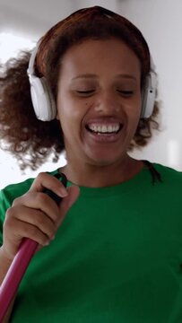 Happy young african american woman having fun singing while cleaning home with broom. Vertical HD screen footage.