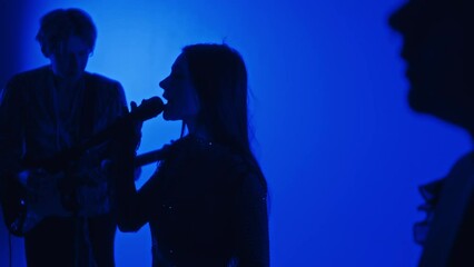 Side footage of silhouettes of female singer and musicians performing music in professional studio with blue colored filter