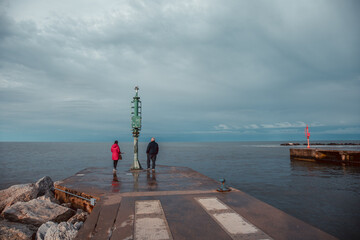 Senior couple having a romantic walk on the pier in a Riviera Romagnola beach location, bright red lady jacket, contrast with the dramatic sky in a winter afternoon. Winter sea.