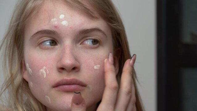 Young woman applying cream foundation on her face using one finger can be useful for creating natural look, helping foundation to melt into skin and making it easy to blend in sheer layer of makeup