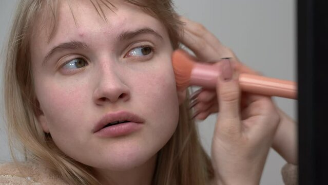 Caucasian female fashion model is applying cream foundation to her face using makeup brush to change natural skin tone. Scene takes place in backstage studio and is captured in handheld shot
