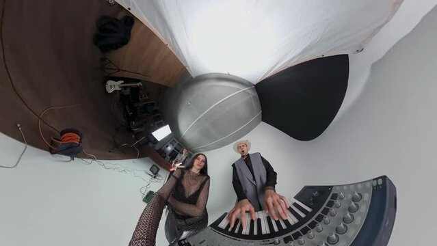 Caucasian blogger and frontwoman of glam rock band filming male keyboardist playing synthesizer using action camera 360 with bassist playing guitar in background in studio